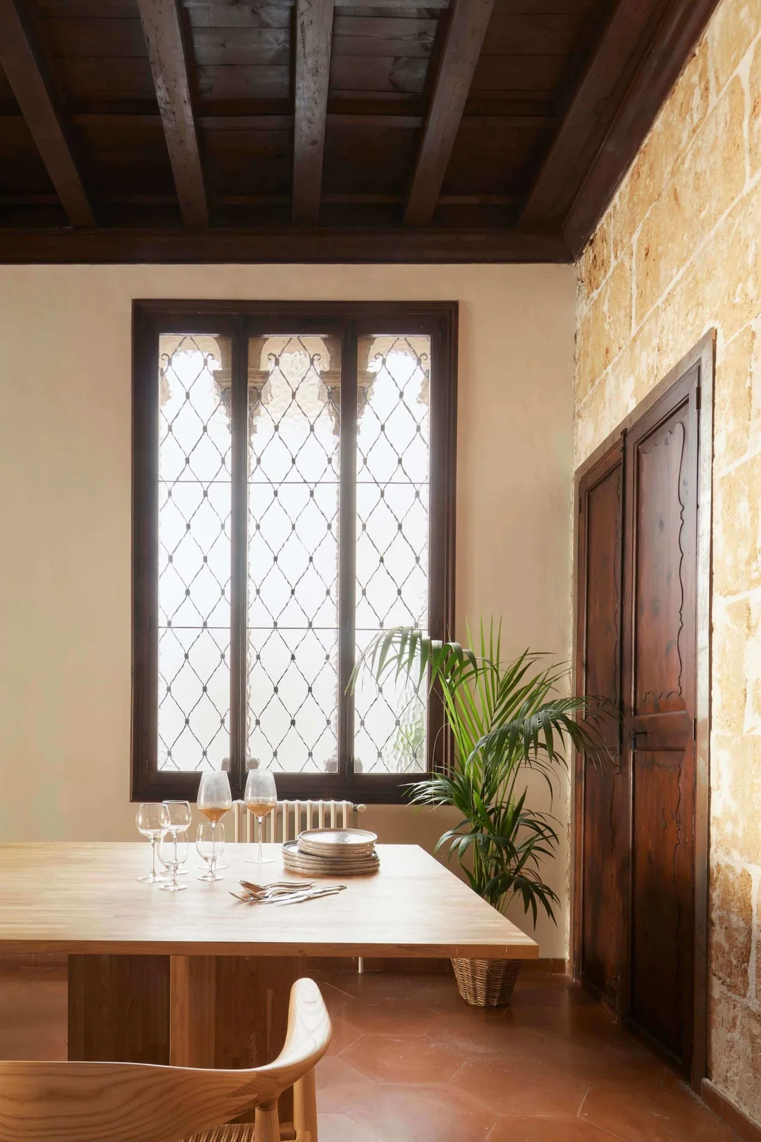 Comedor con mesa de madera junto a una ventana gótica, techo de vigas y muro de marés en una vivienda de Palma.