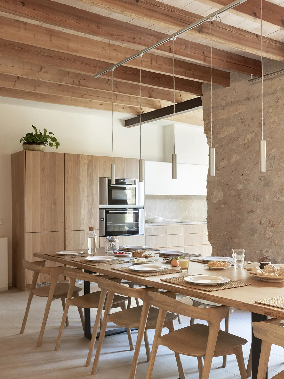 Cocina y comedor bajo vigas de madera, con mesa larga de roble, sillas de madera y lámparas colgantes en Can Cunso.