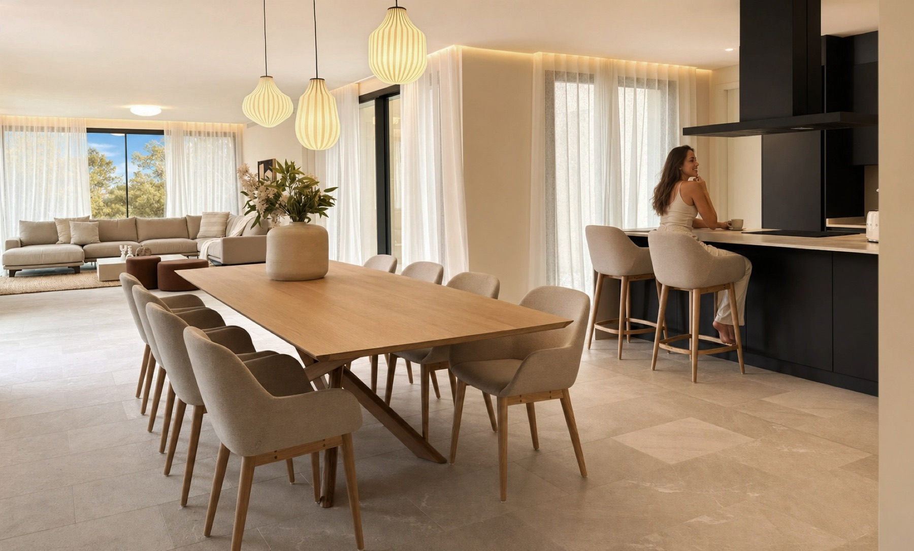 ficha-calavinyes Comedor y cocina de Cala Vinyes: mesa de roble para diez comensales, sillas tapizadas, lámparas de suspensión y una persona apoyada en la barra.