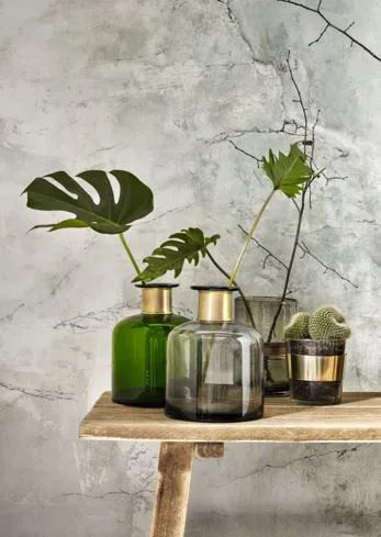 Monstera-Blätter in grünen und grauen Glasvasen auf einem Holzhocker