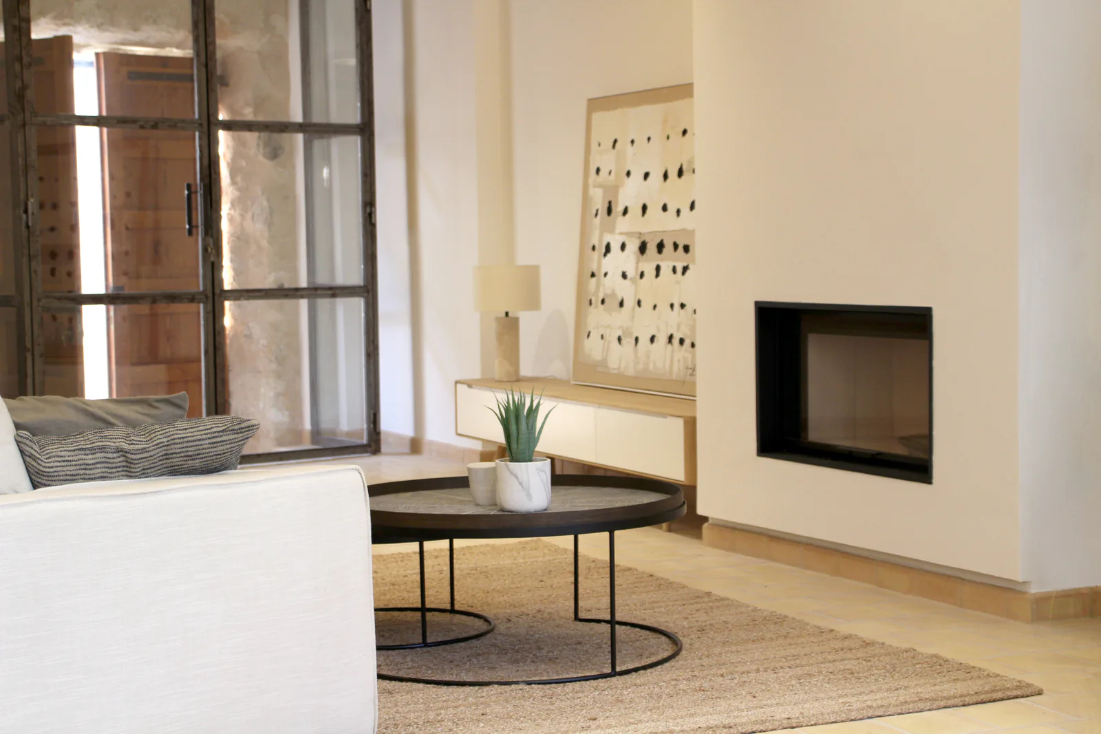Casa de pueblo en Sineu · Home staging Salón de una casa de pueblo en Sineu, con chimenea, alfombra de yute y ventanal de hierro sobre el patio.